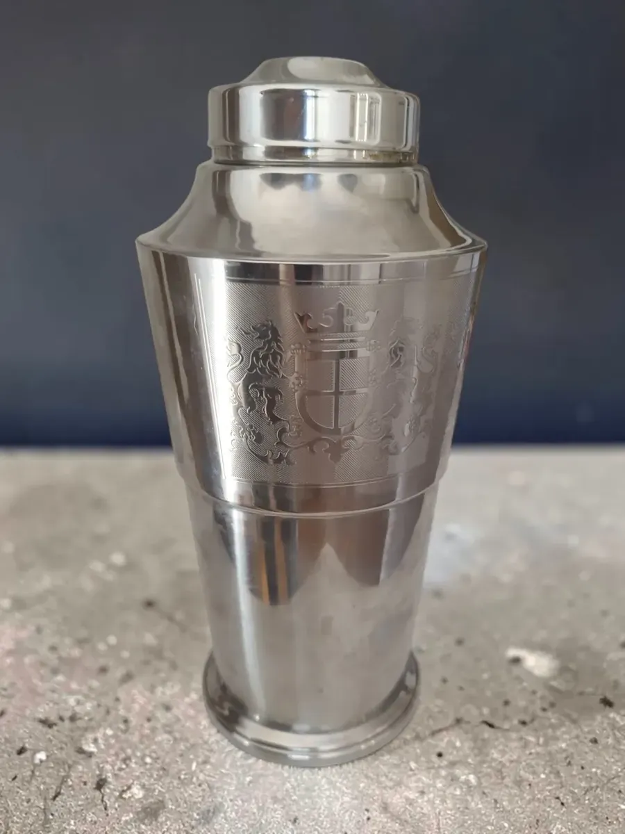 Coctelera de acabado metálico con un escudo grabado, sobre una superficie oscura